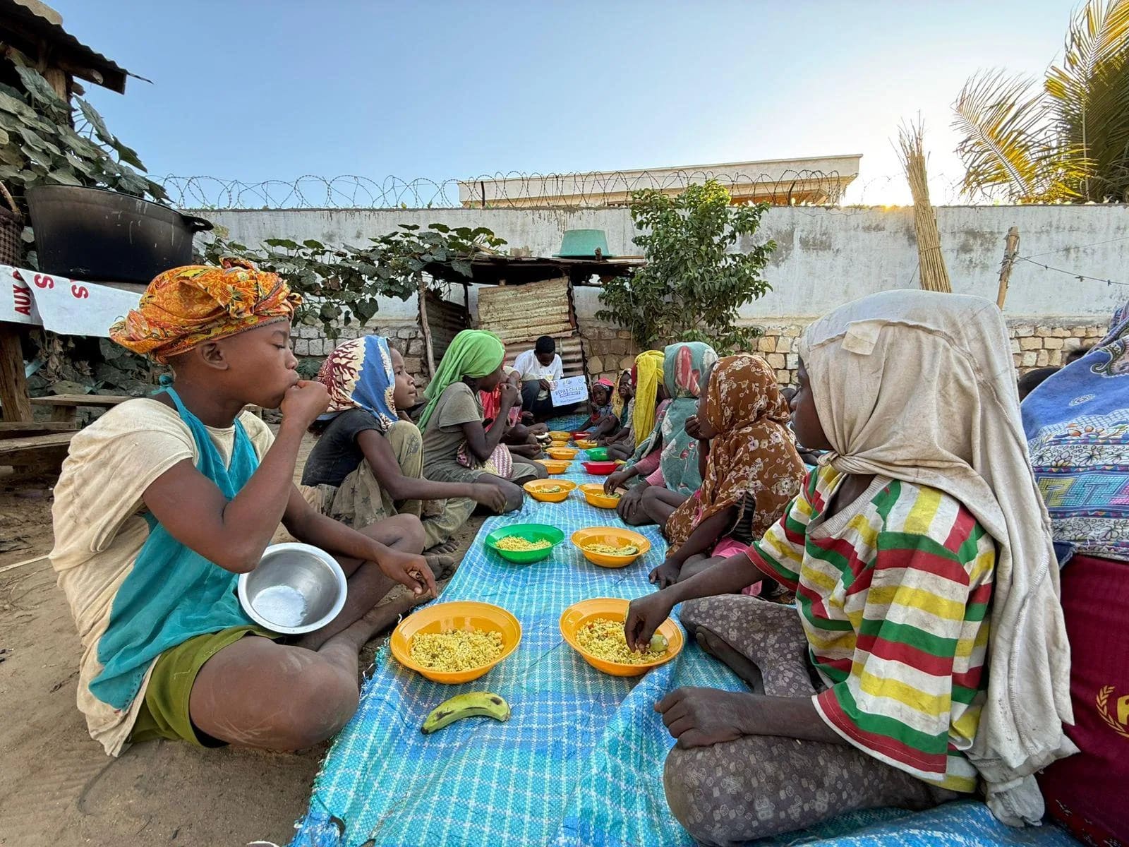 Des enfants mangent un repas chaud, assis en rang sur une natte dans une cour