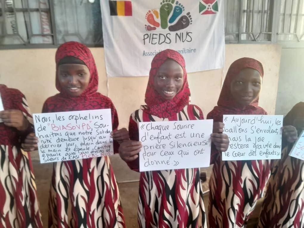 Des filles devant la bannière Pieds Nus aux drapeaux belge et burundais, tenant des messages écrits à la main