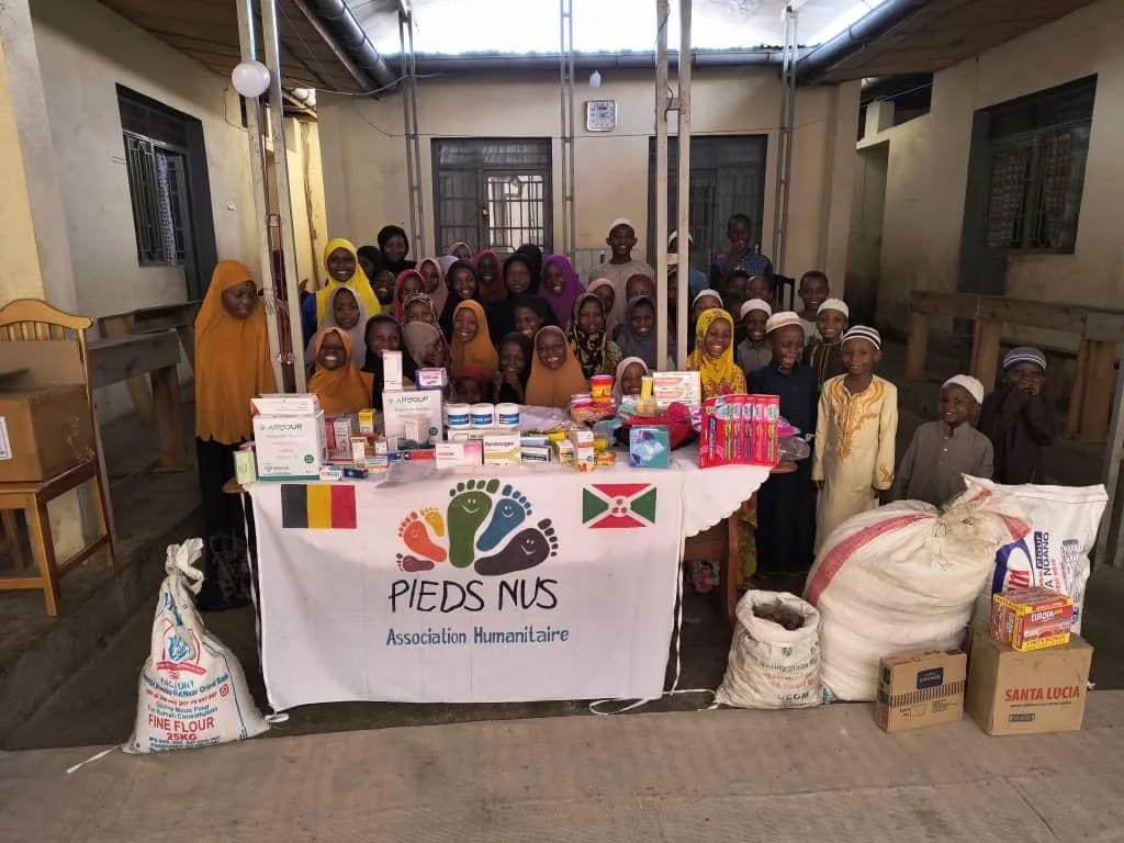 Distribution à Bujumbura : produits, sacs de riz et colis posés devant la bannière Pieds Nus, enfants rassemblés derrière